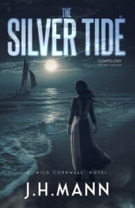 A woman in a long dress stands on a moonlit beach, gazing toward a sailboat on the ocean under a dramatic night sky. The book cover features the title "The Silver Tide" in bold, glowing letters, with the author's name, J.H. Mann, at the bottom and the tagline "A Wild Cornwall Novel" beneath it. A quote from Writing Magazine calls it "Compelling"