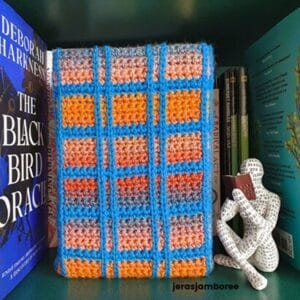 Lennox Crochet Book Cover displayed on a bookshelf in tartan-style stitches of blue, orange, and rust. Alongside are books and a decorative reading figure.