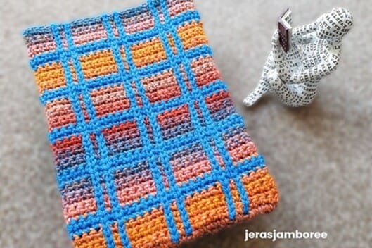 Lennox Crochet Book Cover in blue, orange, and rust tartan design displayed on a carpet beside a decorative reading figure holding a small book.