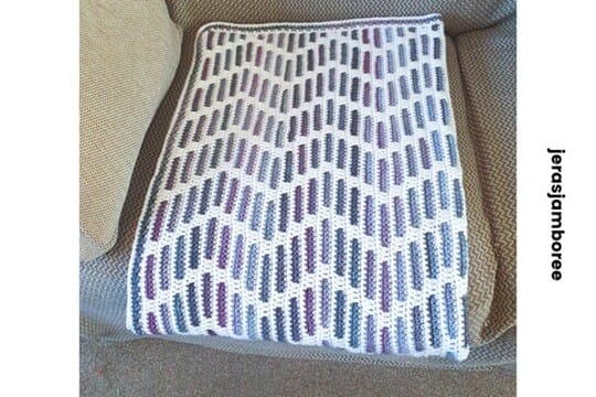 A neatly folded Heather Zig Zag mosaic crochet lap blanket placed on a cushioned armchair. The blanket features a bold zigzag design in purples and blues on a white background, emphasizing the clean stitch work and compact size suitable for a cozy crochet lap blanket.