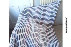A cozy mosaic crochet lap blanket featuring a heather zigzag pattern in purples and blues with a white base, draped over a beige armchair. The repeating geometric design creates a textured, modern look, ideal for showcasing a stylish crochet blanket pattern.