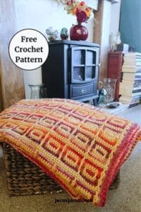 The Amberlight Crochet Blanket draped over a woven basket in front of a fireplace surrounded by books and dried flowers on top of a fire, showing the inset mosaic pattern in warm amber, orange, and purple tones with a textured border. A label reads "Free Crochet Pattern"