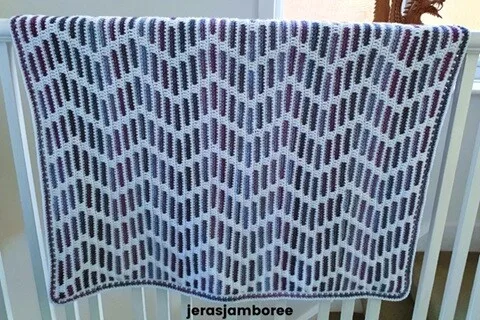 The Heather Zig Zag crochet blanket is draped over a white rail. The pattern features a bold zigzag motif in shades of heather purple and blue against a white background.