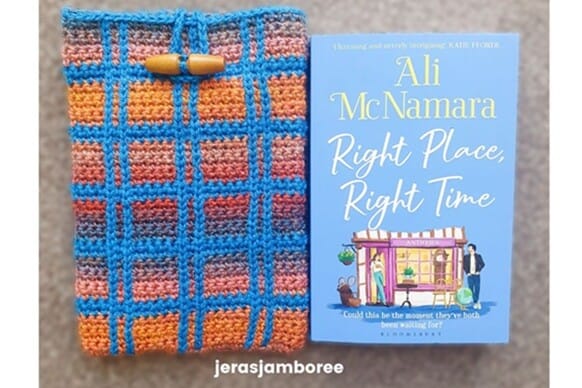 Lennox Crochet Book Sleeve in tartan-style blue, orange, and rust yarn with a wooden toggle closure, shown beside a mass market paperback to show the size on a neutral background.
