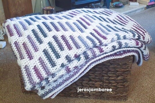 A folded mosaic crochet blanket with a heather zigzag pattern in shades of purple, blue, and white, neatly draped over a woven storage basket. The textured stitches and colorwork highlight the blanket's detailed crochet pattern, making it ideal as a decorative or functional blanket.