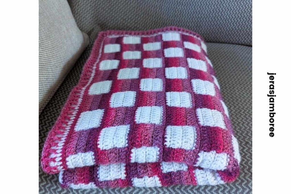 Folded crochet lap blanket with a pink and white gingham pattern resting on a sofa, showing the thickness, texture and finished edge of the Mindful Hands Mosaic Blanket.