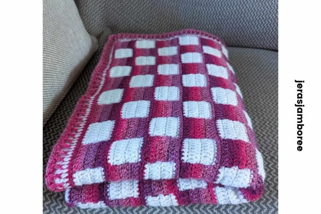 Folded crochet lap blanket with a pink and white gingham pattern resting on a sofa, showing the thickness, texture and finished edge of the Mindful Hands Mosaic Blanket.