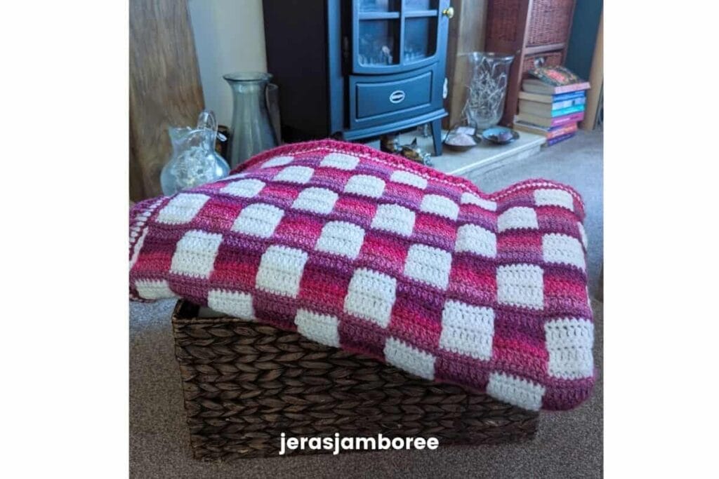Pink and white gingham crochet lap blanket folded over a woven basket, showing the drape and texture of the Mindful Hands Mosaic Blanket in a living room setting.