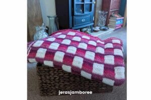 Pink and white gingham crochet lap blanket folded over a woven basket, showing the drape and texture of the Mindful Hands Mosaic Blanket in a living room setting.