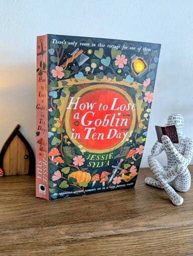 How to Lose a Goblin in Ten Days by Jessie Sylvie standing upright on a wooden surface, with an ornament of a figure reading a book beside it and a fairy-style door in the background.
