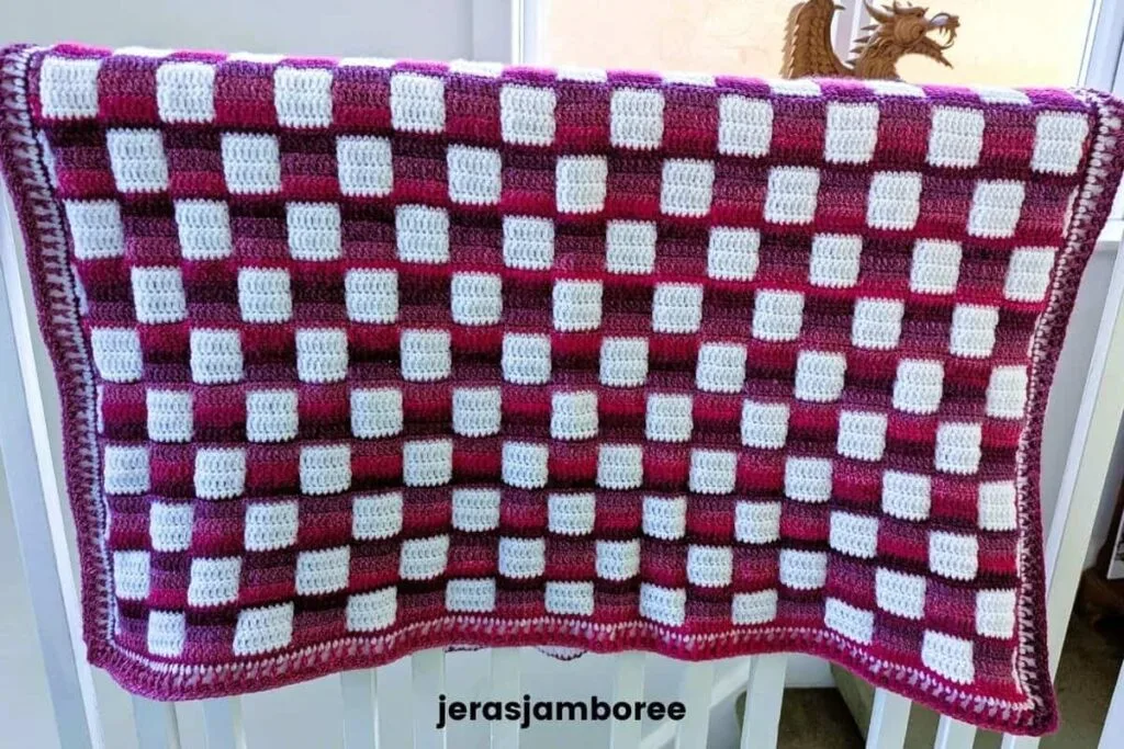 Pink and white gingham crochet lap blanket hanging over a banister, showing the full front of the Mindful Hands Mosaic Blanket and its finished border.