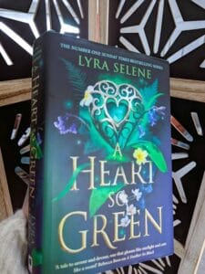 Book cover of A Heart so Green by Lyra Selene, showing a green heart-shaped design with leaves and flowers against a dark background, photographed being held up with a wooden geometrical design in the background.