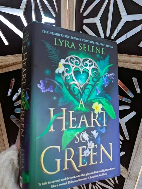 Book cover of A Heart so Green by Lyra Selene, showing a green heart-shaped design with leaves and flowers against a dark background, photographed being held up with a wooden geometrical design in the background.