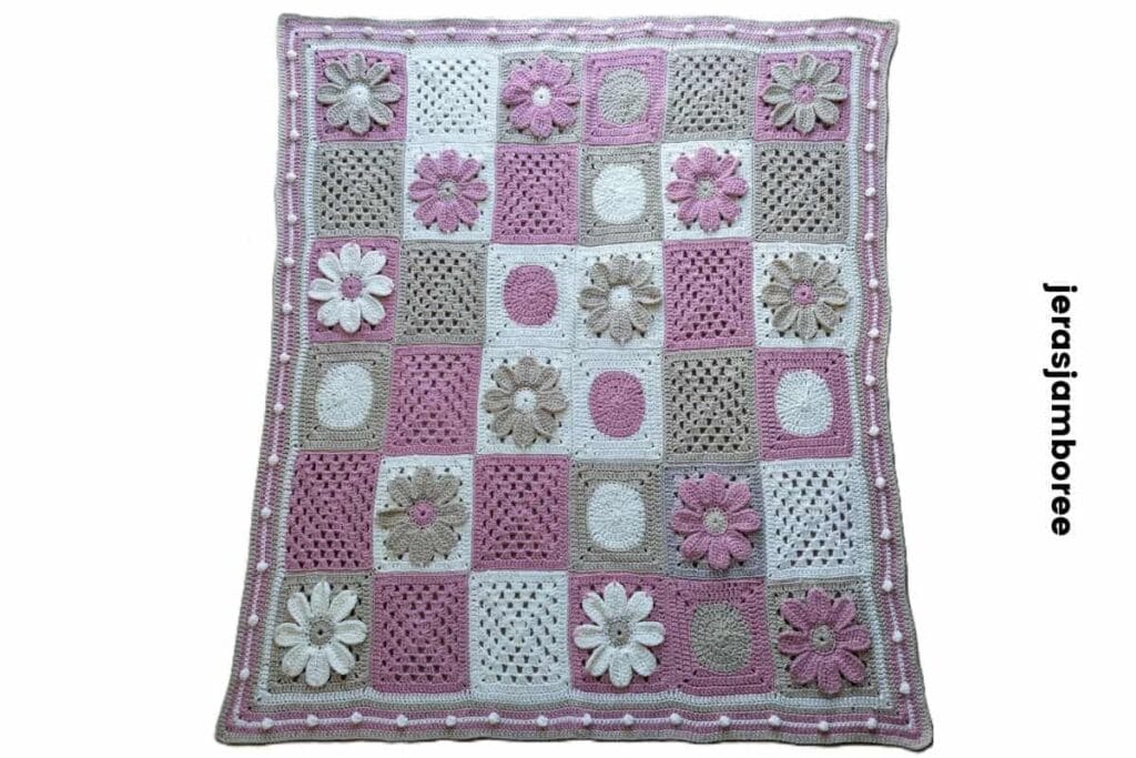 A modern granny square blanket laid flat shows a grid of pink cream and taupe crochet squares including floral motifs and textured stitches arranged evenly across the design. The border for a granny square blanket features a soft pink edge with raised bobble detail framing the blanket and highlighting the finished handmade look.