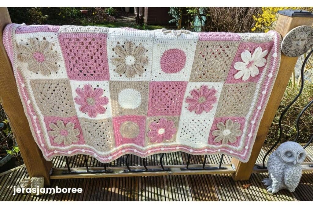 A modern granny square blanket in pink cream and taupe tones is draped over a wooden garden railing in sunlight showing floral and textured crochet squares. The border for a granny square blanket features a soft pink edge with raised bobble detail