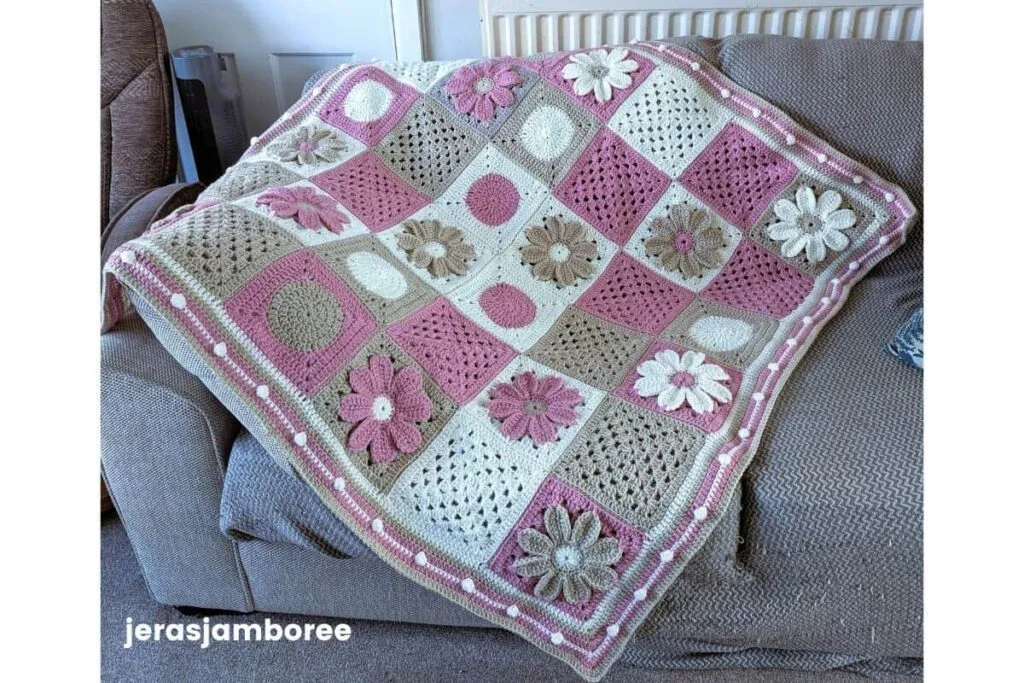 A modern granny square blanket in pink cream and taupe tones is draped over a grey armchair featuring alternating floral and textured crochet squares. The border for a granny square blanket includes a soft pink edge with raised bobble detailing that frames the design and highlights the handmade finish.