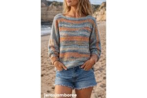 Woman standing on a sandy beach wearing a relaxed striped crochet pullover sweater in blue, grey, and peach tones with balloon sleeves and denim shorts. The photo shows how the finished pullover sweater made from a free crochet pullover pattern looks when worn, with small text "jerasjamboree" at the bottom.