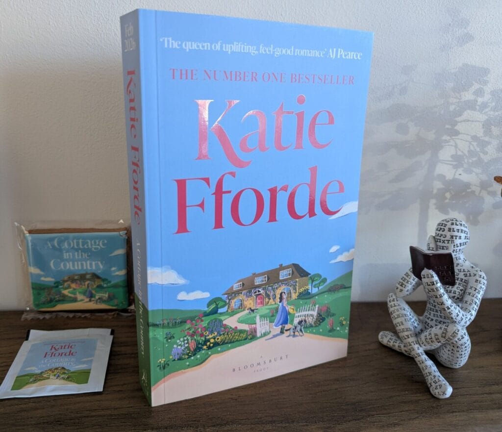 A copy of A Cottage in the Country by Katie Fforde standing upright on a wooden surface, showing the pastel blue cover with a countryside cottage illustration. Beside the book is a small decorative seated figure and matching promotional items including a themed tea bag and small boxed item.