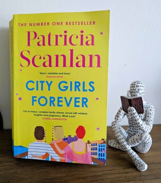 A copy of City Girls Forever by Patricia Scanlan standing upright on a wooden surface, featuring a bright yellow cover with pink and blue title text and an illustration of three women sitting together. Beside the book is a decorative seated reading figure holding a book.