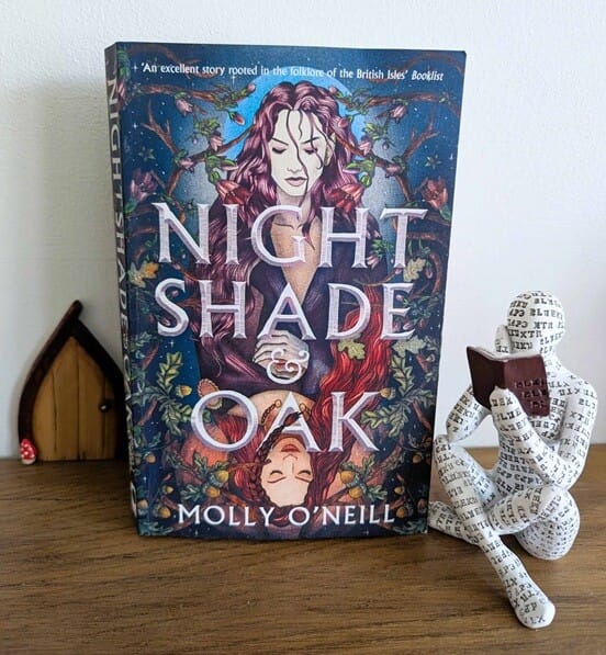 A copy of Nightshade and Oak by Molly O’Neill standing upright on a wooden surface, showing a dark illustrated fantasy cover with mirrored female figures, woodland details, and rich blue and purple tones. Beside the book is a decorative seated reading figure and a small wooden fairy door ornament.