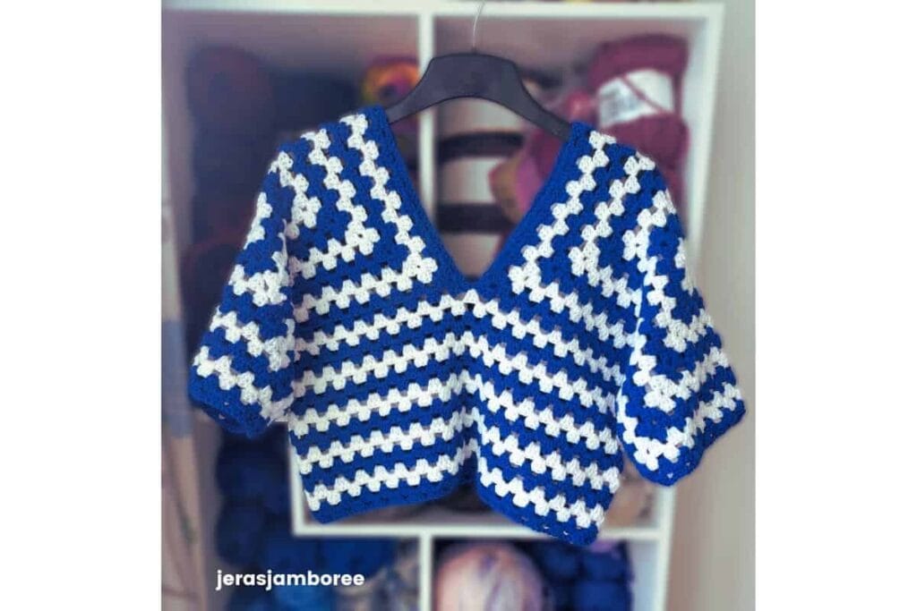 Clover Lane crochet sweater pattern shown in blue and white, hanging in front of a yarn of shelves