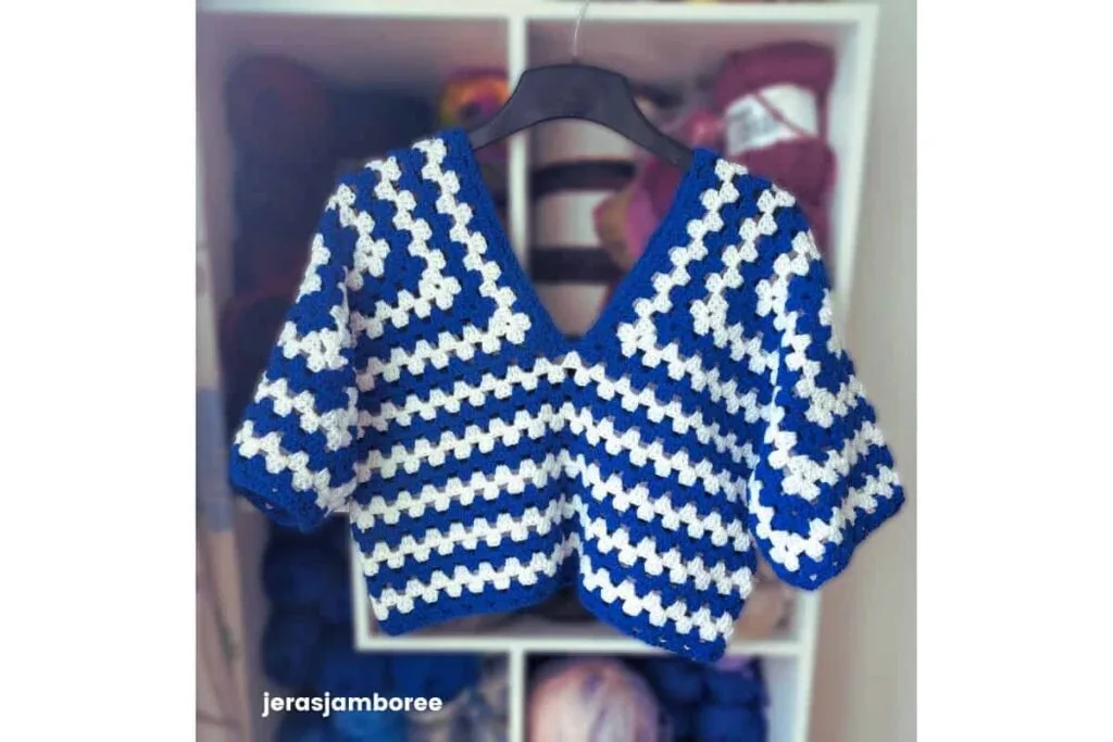 Clover Lane crochet sweater pattern shown in blue and white, hanging in front of shelves of yarn