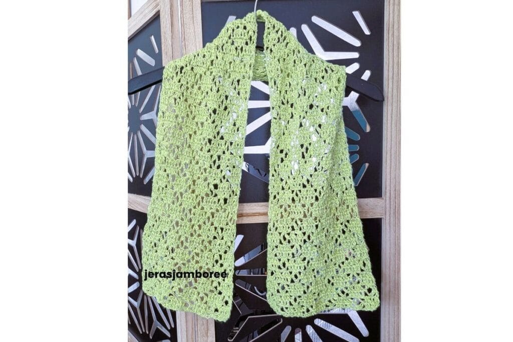 Lightweight lime green crochet lace scarf hanging on a black hanger against a decorative screen, showing the airy openwork stitch pattern and drape of the Meadowsong scarf design.