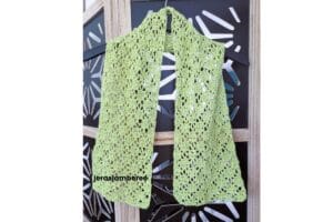 Lightweight lime green crochet lace scarf hanging on a black hanger against a decorative screen, showing the airy openwork stitch pattern and drape of the Meadowsong scarf design.