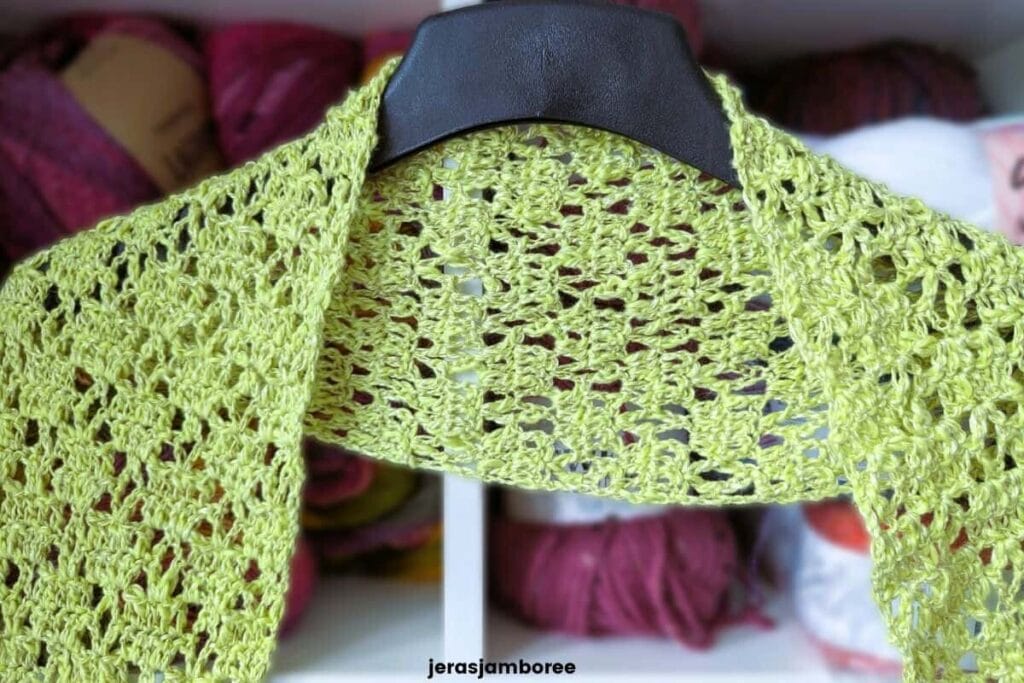 Close-up of the lightweight Meadowsong crochet scarf draped over a black hanger, showing the lacy openwork stitch pattern with colourful yarn skeins blurred in the background.