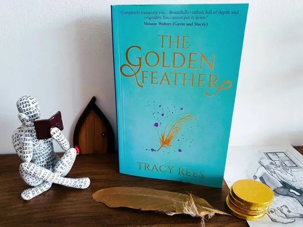 A copy of The Golden Feather by Tracy Rees standing on a wooden surface beside a decorative seated figure holding a small book. In front of the novel is a large golden feather, with stacked gold coins and an illustrated page to the side. The turquoise book cover features gold lettering and a feather illustration, creating a magical fairytale atmosphere.