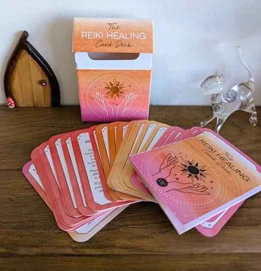 A spread of pink and orange Reiki Healing cards displayed on a wooden surface beside the matching card deck box. The cards feature hand illustrations and spiritual symbols, with a small glass cat ornament and wooden decorative fairy door in the background.