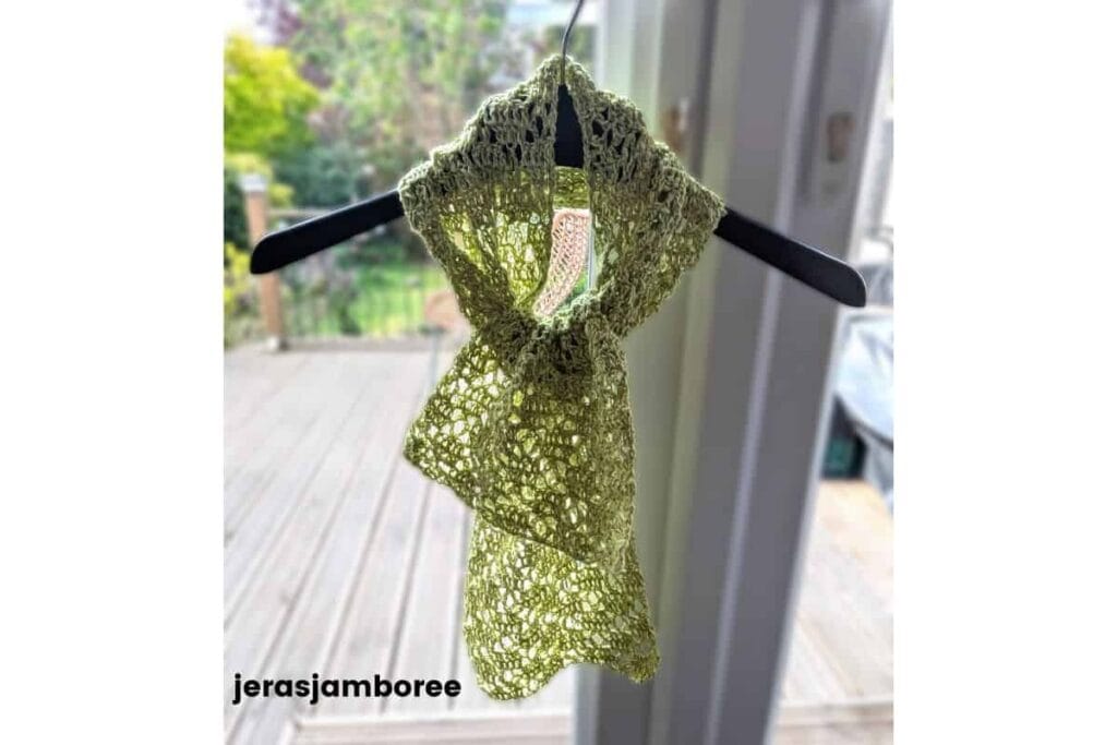 Lightweight lime green crochet lace scarf tied loosely on a hanger in front of a softly blurred garden background, showing the airy open stitch pattern and drape of the Meadowsong scarf.