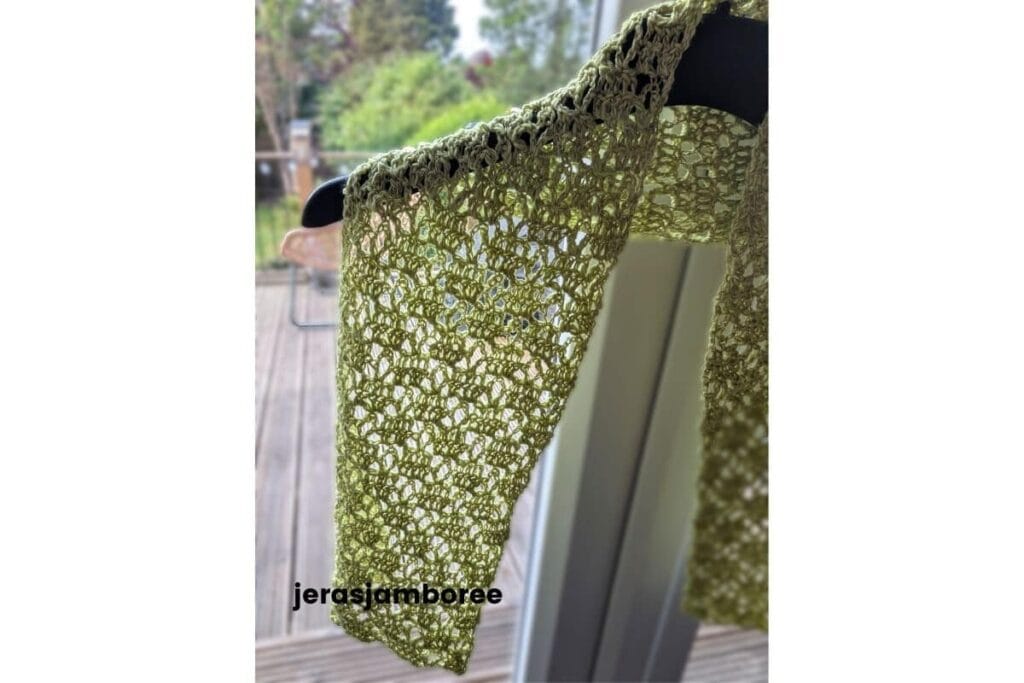 Close-up of the lightweight Meadowsong crochet scarf hanging on a black hanger, showing the airy lace stitch pattern and soft drape against a blurred garden background.