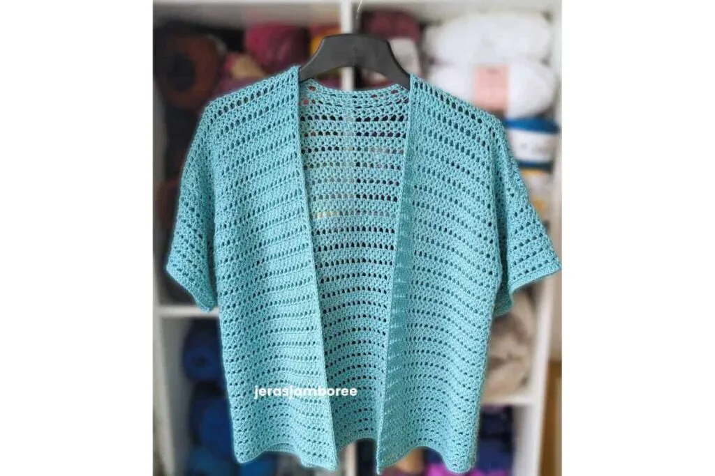 Sandrine short sleeve crochet cardigan in aqua blue featuring an open front design and lightweight mesh stitch hanging in front of a yarn shelf