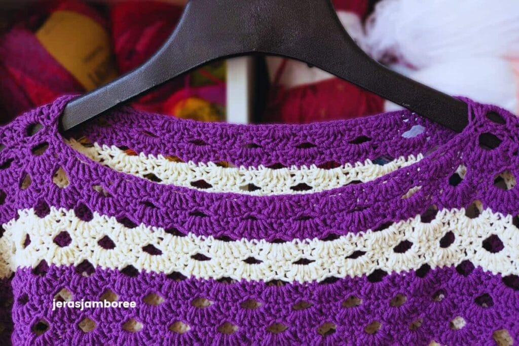 Close-up of the boat-neck neckline on the Coralie crochet poncho, showing the purple and cream graduated stripes and the open Arcade stitch pattern worked in a lightweight cotton blend yarn.