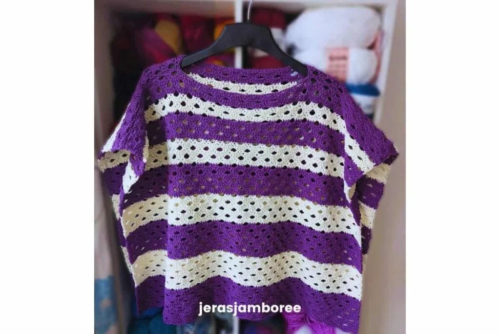 Coralie crochet poncho hanging on a black hanger, worked in alternating purple and cream graduated stripes using the Arcade stitch. This lightweight crochet poncho features a relaxed boat-neck neckline, airy fabric, and an easy two-panel construction suitable for spring and summer layering.