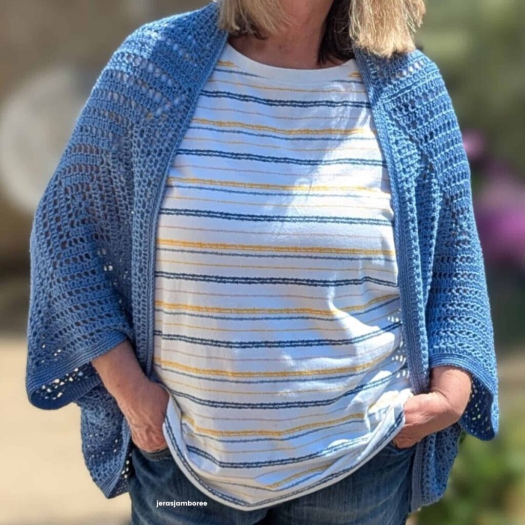 Close-up of the Aveline Granny Square Shrug worn over a striped top, showing the open-front cocoon shape, oversized granny square construction and lightweight drape of the blue crochet fabric.