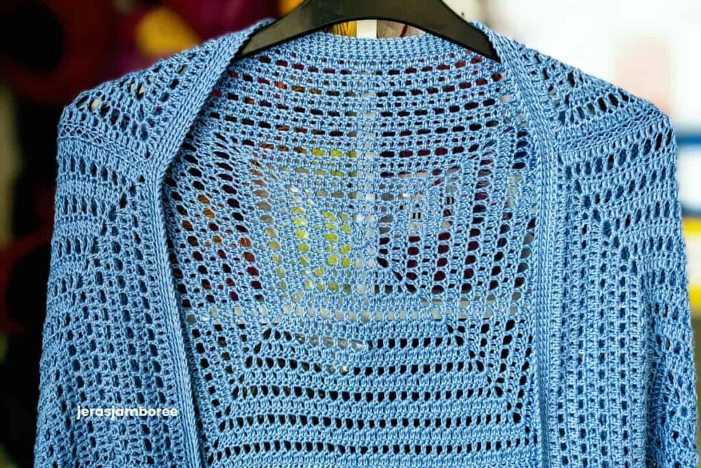 Close-up front view of the Aveline Cocoon Cardigan hanging on a hanger, highlighting the central oversized granny square, open crochet stitch pattern and neatly finished neckline in soft blue DK bamboo yarn.