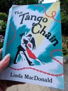 The Tango Chain by Linda MacDonald paperback book cover held outdoors in a garden.