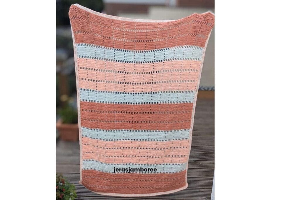 Marlowe crochet lap blanket in peach, off white and dusky peach with geometric filet blocks and open mesh rows is being held up to show the full length and width