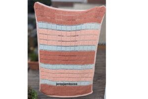 Marlowe crochet lap blanket in peach, off white and dusky peach with geometric filet blocks and open mesh rows is being held up to show the full length and width