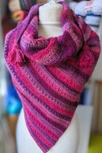 Mariselle crochet shawl wrapped around a mannequin, showing the pink and purple self-striping yarn, textured fabric and tassel details