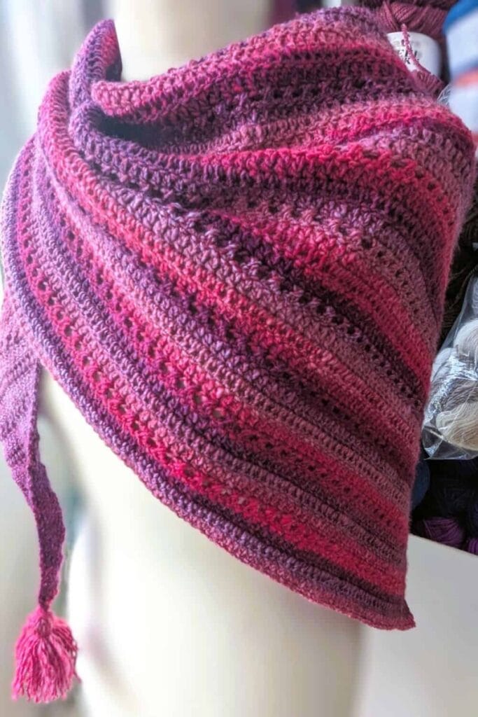 Mariselle crochet shawl draped loosely over a mannequin, showing the diagonal direction of the textured pink and purple fabric and tassel at the front point