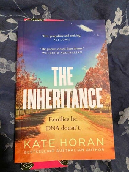 Book cover of The Inheritance by Kate Horan, featuring a red dirt road lined with autumn-coloured trees beneath a blue and purple sky. The tagline reads, “Families lie. DNA doesn’t.”