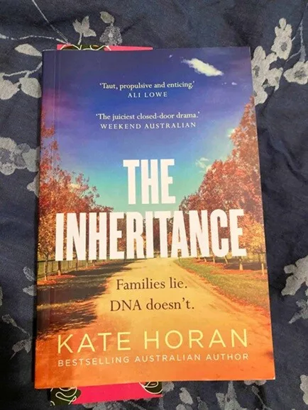 Book cover of The Inheritance by Kate Horan, featuring a red dirt road lined with autumn-coloured trees beneath a blue and purple sky. The tagline reads, “Families lie. DNA doesn’t.”
